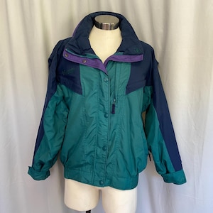 May include: A vintage teal, navy, and purple jacket. The jacket features a high collar, button and zipper closure, and a color-blocked design. The sleeves are navy, and the jacket is displayed on a mannequin.