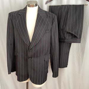 May include: A dark gray pinstripe suit, including a blazer and matching trousers, displayed on a mannequin. The blazer has a two-button closure and a notched lapel. The pinstripes are a combination of red and blue. The trousers are neatly folded.