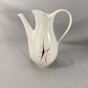 May include: Cream-colored ceramic pitcher with a long spout and curved handle. The pitcher features a mid-century modern design with a black and pink abstract starburst pattern. A decorative piece for serving beverages.