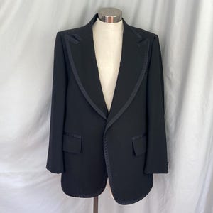 May include: A black tuxedo jacket featuring a satin shawl lapel, two flap pockets with satin trim, and a single-button closure. The jacket is displayed on a mannequin, showcasing long sleeves and a structured, tailored fit with clean lines.