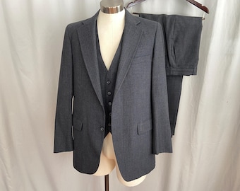 May include: A three-piece charcoal gray suit displayed on a mannequin. The suit includes a blazer, vest, and trousers. The blazer has a notched lapel and two front pockets. The vest has a button-down closure. The trousers are folded and hung on a wooden hanger.