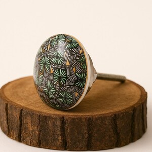 Pode incluir: Um puxador decorativo para armário com um design redondo e estampado. O puxador apresenta um padrão geométrico preto, branco e verde com detalhes dourados. O puxador é montado numa base e parafuso prateados, e é exibido numa superfície de madeira.