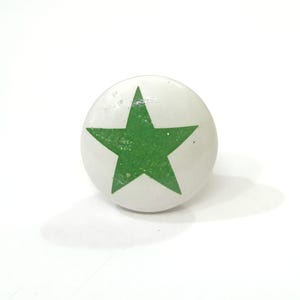 May include: A white, round cabinet knob featuring a green star design. The star is centrally placed and has a textured finish. The knob is probably made of ceramic or a similar material.