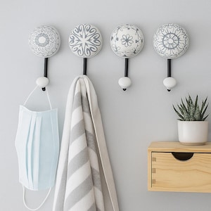 May include: Four decorative wall hooks with white and grey patterned ceramic knobs. Each hook has a black stem and a white base. A grey and white striped towel and a blue face mask hang from two of the hooks. A small potted plant and a wooden drawer are also visible.