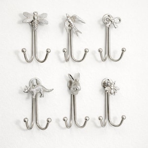 May include: Six silver-colored metal wall hooks with decorative animal and object designs. The hooks feature a dragonfly, bird, bow, dinosaur, rabbit, and bee. Each hook has two lower prongs for hanging items.