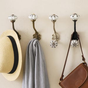 May include: Four decorative wall hooks with white ceramic knobs and ornate metal arms. A straw hat with a black band, a gray scarf, and a brown leather bag hang from the hooks. The hooks are mounted on a light beige wall.