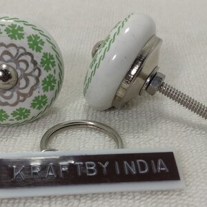 May include: Two white ceramic knobs with green and silver floral designs. The knobs have a metal screw attachment. A white tag with the text "KRAFTBY INDIA" is included.