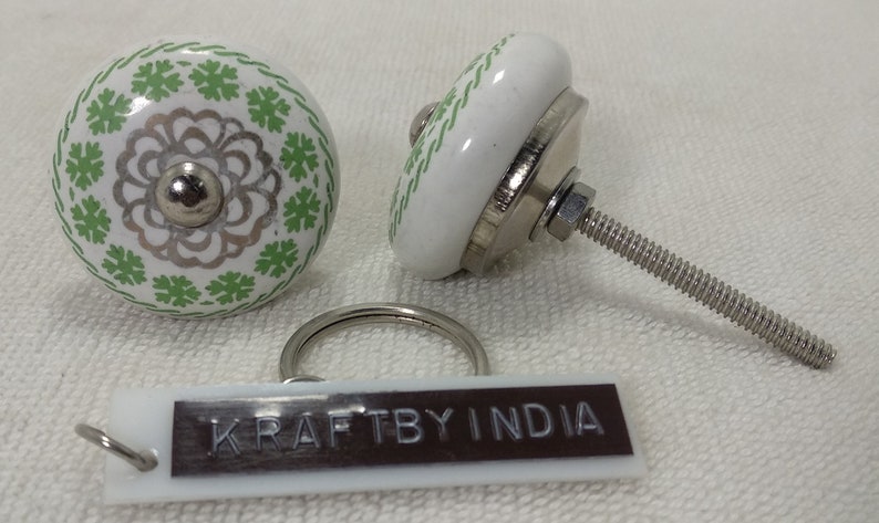 May include: Two white ceramic knobs with green and silver floral designs. The knobs have a metal screw attachment. A white tag with the text "KRAFTBY INDIA" is included.