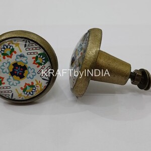 May include: Two round cabinet knobs with a brass finish. The knobs have a white background with a colorful floral pattern. One knob is attached to a screw.