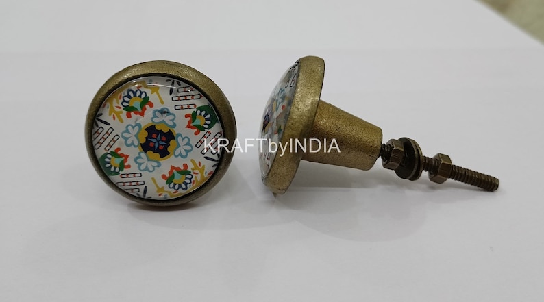 May include: Two round cabinet knobs with a brass finish. The knobs have a white background with a colorful floral pattern. One knob is attached to a screw.