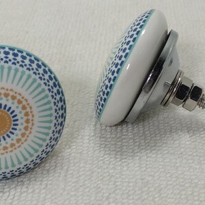 May include: Two ceramic cabinet knobs with a blue, white, and orange geometric pattern. The knobs have a silver metal base and a screw for attaching to a cabinet door.