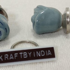 May include: Two blue ceramic rose-shaped drawer knobs with silver hardware. The knobs are attached to a white tag with the text "KRAFTBY INDIA".