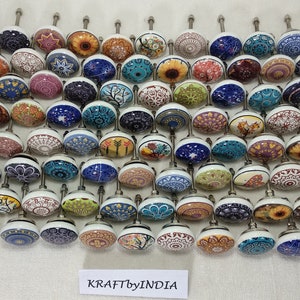 May include: A collection of 50 colourful ceramic cabinet knobs with various patterns and designs. The knobs are round and have a silver metal base. The knobs are arranged in rows on a white surface. KRAFT by INDIA is printed on the surface.