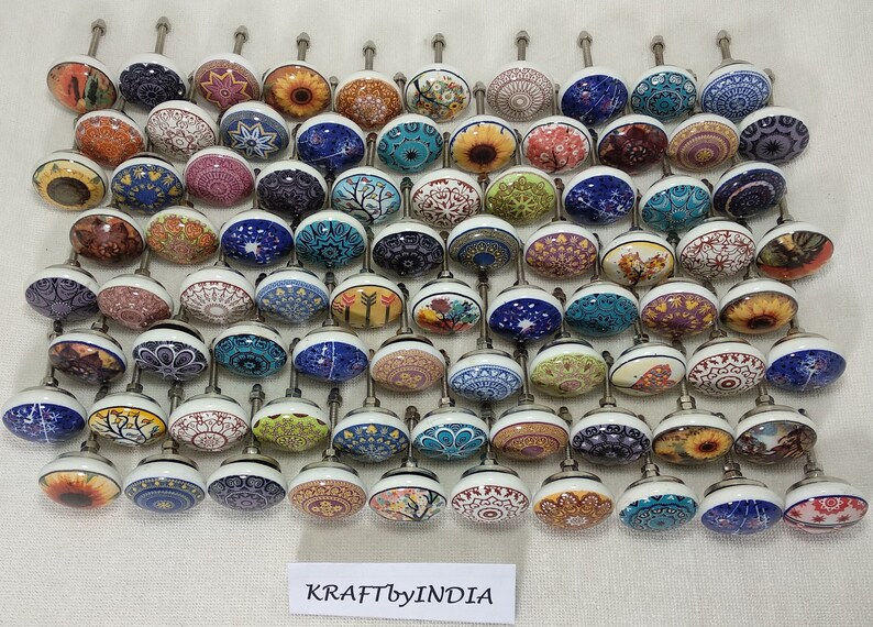May include: A collection of 50 colourful ceramic cabinet knobs with various patterns and designs. The knobs are round and have a silver metal base. The knobs are arranged in rows on a white surface. KRAFT by INDIA is printed on the surface.