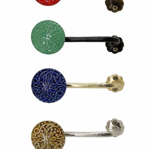 Puede incluir: Cuatro ganchos de pared decorativos con pomos redondos de cer&aacute;mica texturizada en rojo, verde, azul y beige. Cada gancho tiene un brazo de metal curvado y una base decorativa en forma de flor. Ideales para colgar objetos.