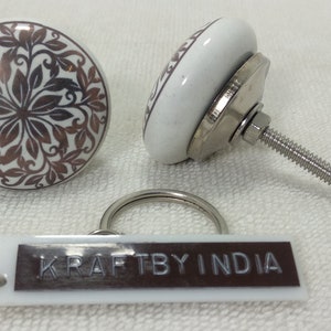 May include: Two white ceramic knobs with a brown floral design and a silver metal screw. A white plastic keychain with the text "KRAFTBY INDIA" in brown.