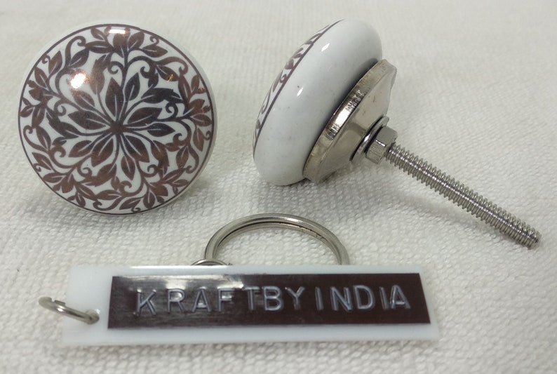 May include: Two white ceramic knobs with a brown floral design and a silver metal screw. A white plastic keychain with the text "KRAFTBY INDIA" in brown.