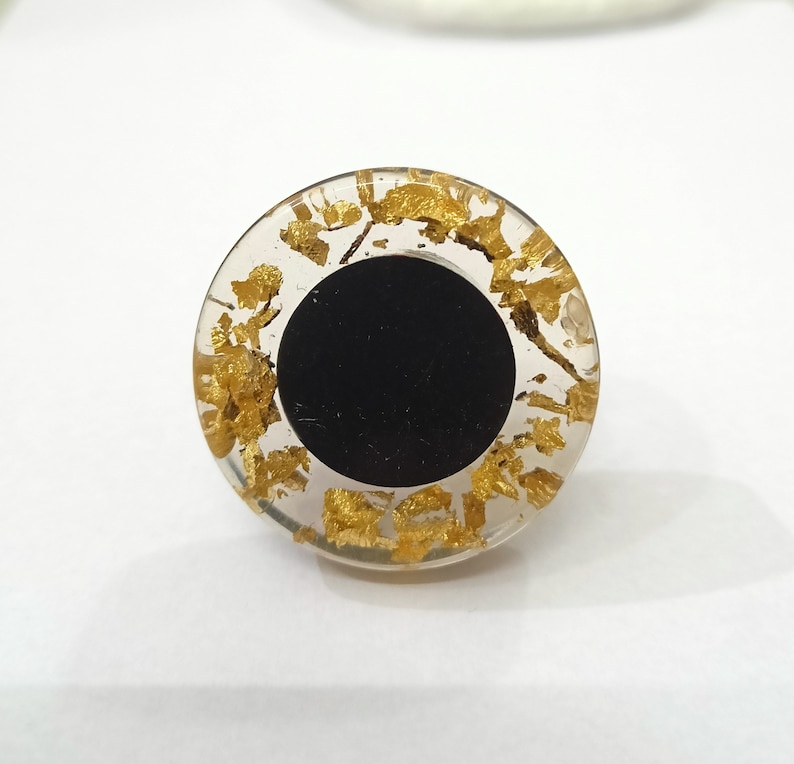 Puede incluir: Un anillo de resina transparente con un centro negro y escamas doradas incrustadas en la resina.