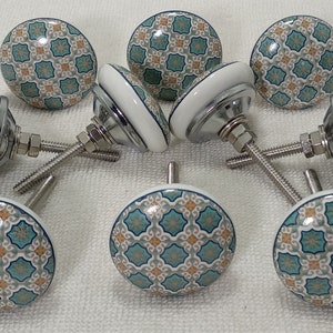 以下が含まれることがあります： 白いベースとターコイズブルーとベージュの幾何学模様が施されたセラミック製のキャビネットノブ8個セット。各ノブには銀色のネジが付いています。
