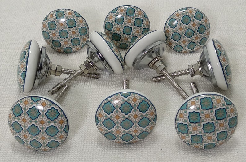 以下が含まれることがあります： 白いベースとターコイズブルーとベージュの幾何学模様が施されたセラミック製のキャビネットノブ8個セット。各ノブには銀色のネジが付いています。