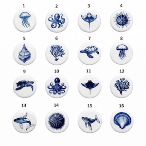 May include: A collection of sixteen white round ceramic buttons with blue sea creature designs. The designs include a jellyfish, octopus, stingray, sea urchin, seashell, coral, sea turtle, and whale. The buttons are arranged in four rows of four.