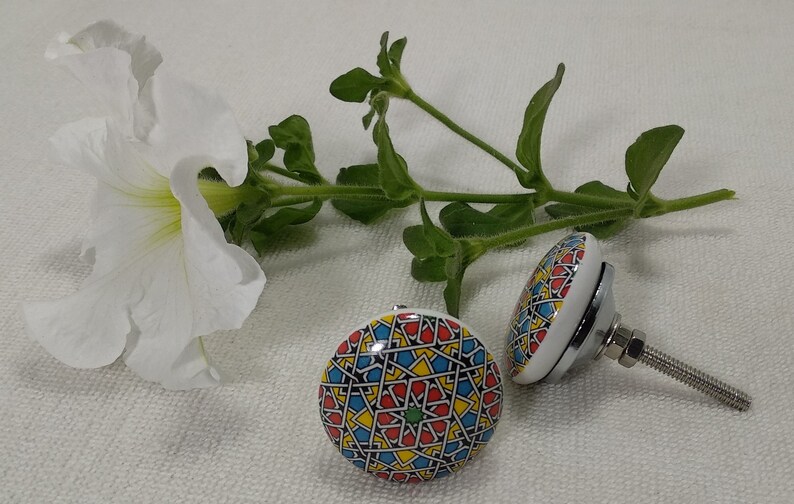 May include: Two ceramic cabinet knobs with a colourful geometric pattern in shades of red, yellow, blue, and green. One knob is attached to a silver screw.