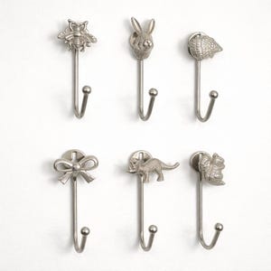 May include: Six silver-colored metal wall hooks. Each hook features a different decorative top: an owl, a rabbit, a seashell, a bow, a dinosaur, and a squirrel. The hooks are designed for hanging items.