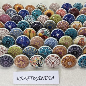 May include: A collection of 50 colourful ceramic cabinet knobs with various patterns and designs. The knobs are round and have a white base. The patterns include floral, geometric, and abstract designs. The knobs are arranged in a grid pattern on a white surface. The text "KRAFTbyINDIA" is visible at the bottom of the image.