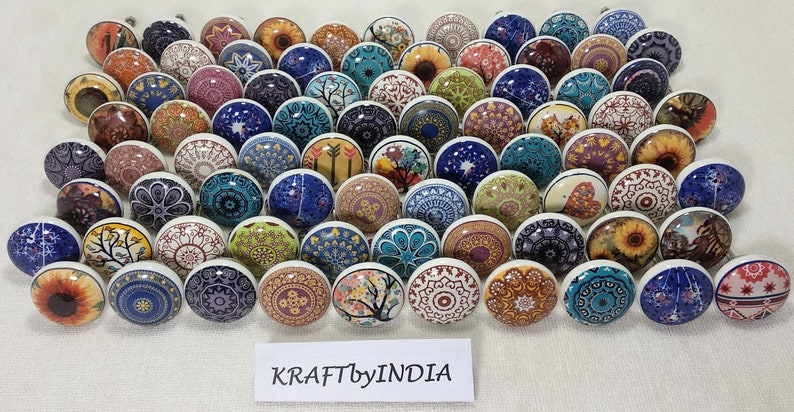 May include: A collection of 50 colourful ceramic cabinet knobs with various patterns and designs. The knobs are round and have a white base. The patterns include floral, geometric, and abstract designs. The knobs are arranged in a grid pattern on a white surface. The text "KRAFTbyINDIA" is visible at the bottom of the image.