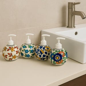 Puede incluir: Cuatro dispensadores de jabón de cerámica con tapas de bomba blancas. Cada dispensador presenta un patrón floral diferente en varios colores, incluyendo rojo, amarillo, azul y verde, sobre un fondo blanco. Los dispensadores están colocados sobre una encimera blanca cerca de un lavabo.
