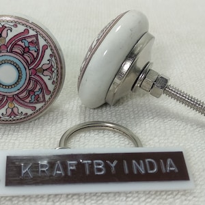 May include: Two white ceramic cabinet knobs with a floral design in pink, blue, and gold. The knobs have a metal base and screw. A small tag with the text "KRAFTBY INDIA" is also present.