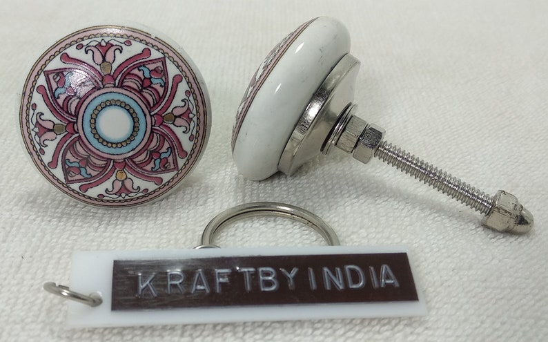 May include: Two white ceramic cabinet knobs with a floral design in pink, blue, and gold. The knobs have a metal base and screw. A small tag with the text "KRAFTBY INDIA" is also present.