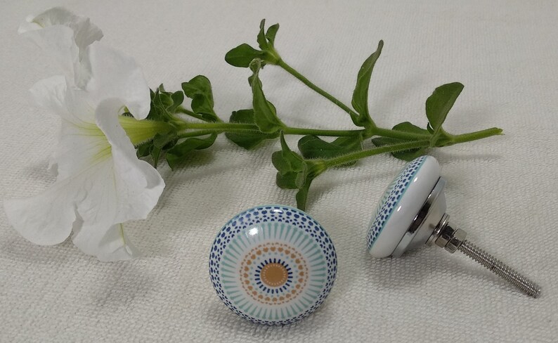 May include: Two white ceramic cabinet knobs with a blue and orange geometric pattern. The knobs have a silver screw.