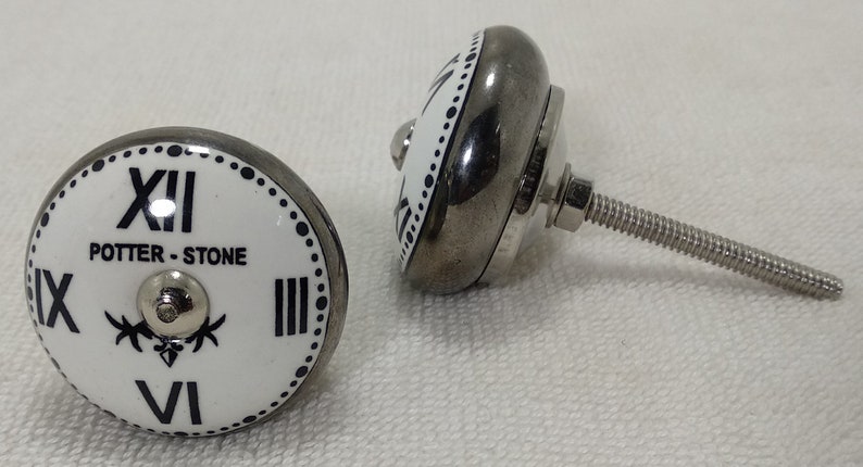 May include: Two white ceramic cabinet knobs with a black clock face design. The knobs have a silver metal base and a screw for attaching to a cabinet door. The clock face features Roman numerals and the text "POTTER - STONE".