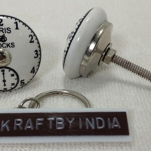 May include: White ceramic knob with a black clock face design. The clock face reads "Paris Clocks" and has black numbers and hands. The knob has a silver metal screw and a silver metal base. The knob is attached to a white and brown keychain with the text "Kraftby India".
