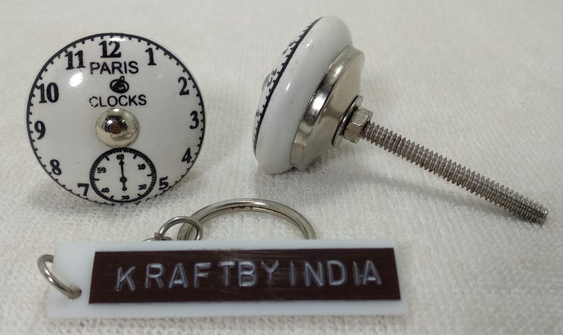 May include: White ceramic knob with a black clock face design. The clock face reads "Paris Clocks" and has black numbers and hands. The knob has a silver metal screw and a silver metal base. The knob is attached to a white and brown keychain with the text "Kraftby India".
