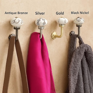 May include: Four decorative wall hooks in antique bronze, silver, gold, and black nickel finishes. Each hook has a white ceramic knob with a decorative pattern. A brown strap, a pink scarf, and a grey towel hang from the hooks.