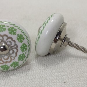 May include: Two white ceramic cabinet knobs with a green and silver floral design. One knob has a screw attached.