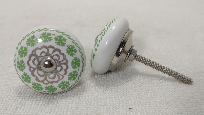 May include: Two white ceramic cabinet knobs with a green and silver floral design. One knob has a screw attached.