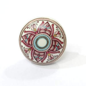 May include: A decorative ceramic cabinet knob with a floral design. The knob features a white base with a central design in shades of pink, blue, and gold. The floral pattern is symmetrical and detailed, with a light pink border.