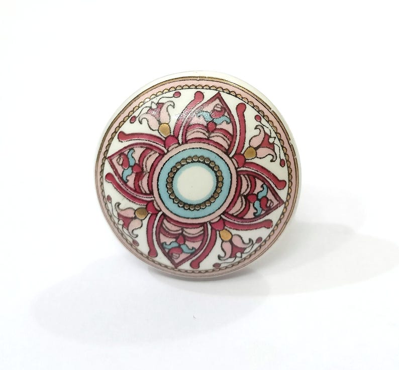 May include: A decorative ceramic cabinet knob with a floral design. The knob features a white base with a central design in shades of pink, blue, and gold. The floral pattern is symmetrical and detailed, with a light pink border.