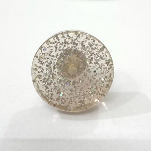 Puede incluir: Un pomo decorativo redondo y transparente con purpurina plateada incrustada en la resina. El pomo tiene una parte superior plana y una parte inferior redondeada.