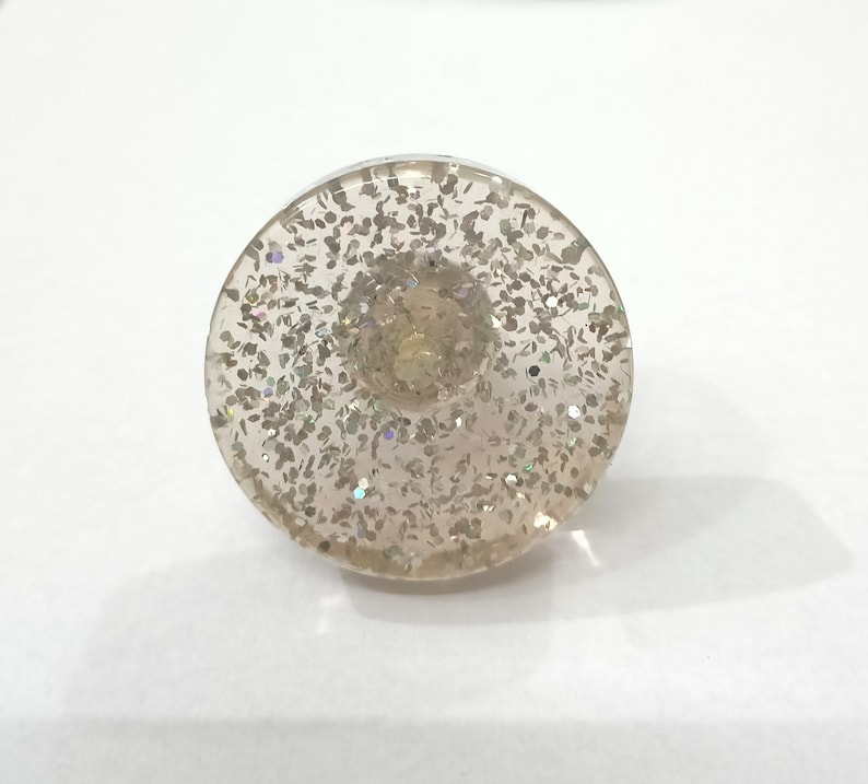 Puede incluir: Un pomo decorativo redondo y transparente con purpurina plateada incrustada en la resina. El pomo tiene una parte superior plana y una parte inferior redondeada.
