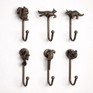 May include: Six bronze-colored metal wall hooks with animal designs: a squirrel, fox, dinosaur, seahorse, parrot, and bear. Each hook has a decorative animal head and a curved hook for hanging items.