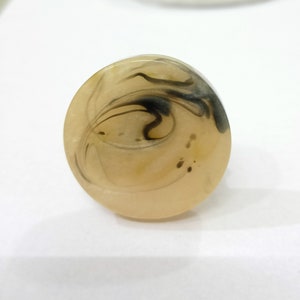 Puede incluir: Un anillo ovalado beige con patrones negros en espiral en el interior. El anillo est&aacute; hecho de resina y tiene un acabado liso y brillante.