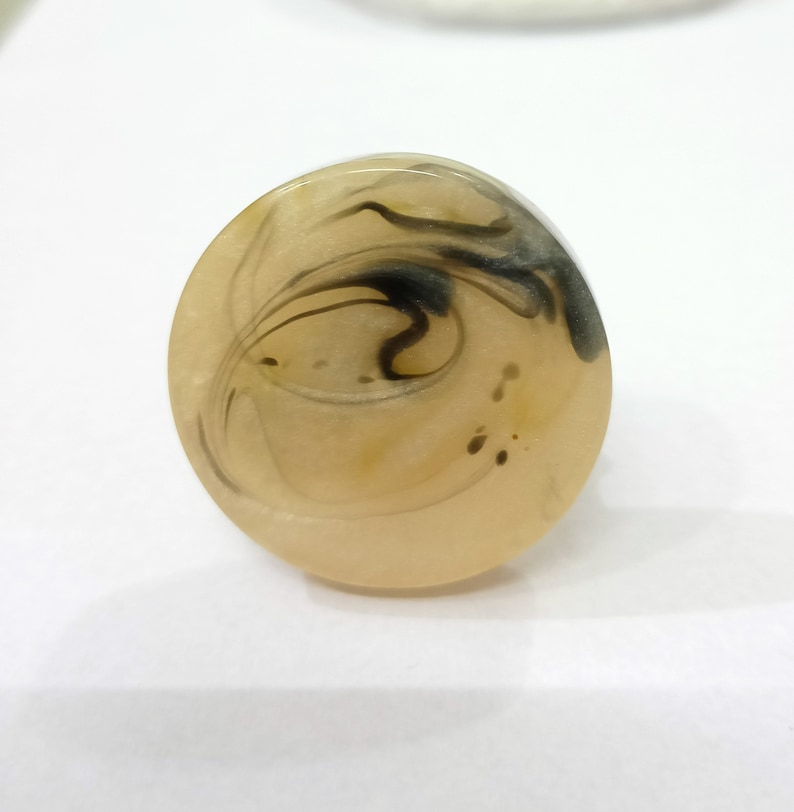 Puede incluir: Un anillo ovalado beige con patrones negros en espiral en el interior. El anillo est&aacute; hecho de resina y tiene un acabado liso y brillante.