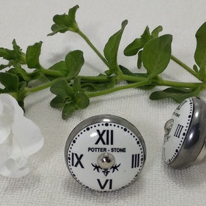 May include: Two white ceramic knobs with a black clock face design. The knobs have a silver metal base and a screw for attaching to a surface. The clock face features Roman numerals and the text "POTTER - STONE".