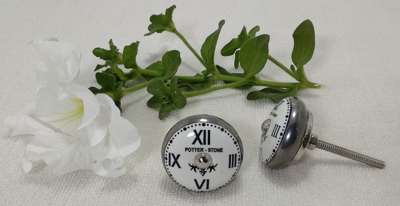 May include: Two white ceramic knobs with a black clock face design. The knobs have a silver metal base and a screw for attaching to a surface. The clock face features Roman numerals and the text "POTTER - STONE".