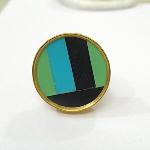 Puede incluir: Un anillo redondo de color dorado con un dise&ntilde;o geom&eacute;trico negro, verde y azul.