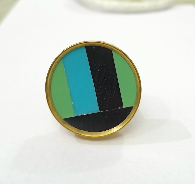 Puede incluir: Un anillo redondo de color dorado con un dise&ntilde;o geom&eacute;trico negro, verde y azul.
